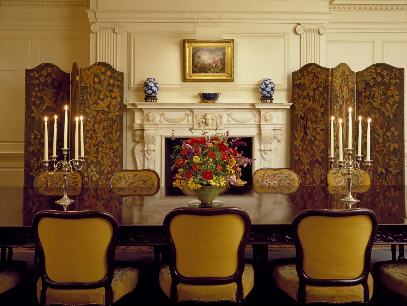 A polished dining table with lit silver candelabra and a bowl of red and yellow flowers before a carved mantel and embroidered screens