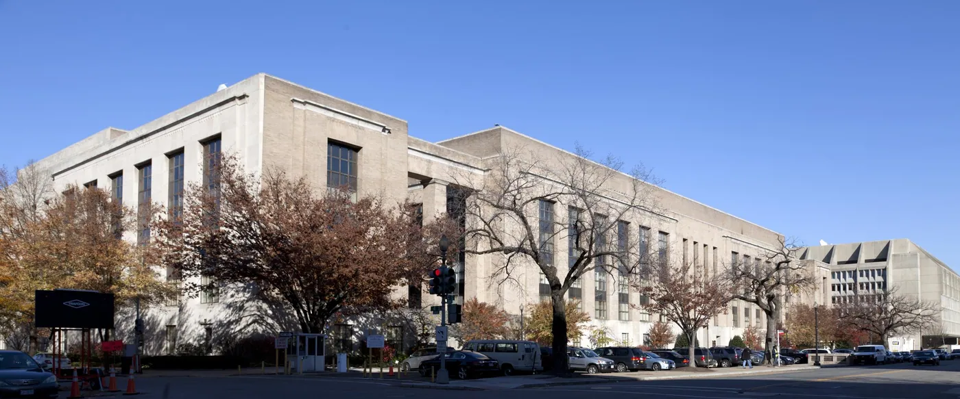 A long, pale limestone federal building of stripped-classical design under a clear blue sky, with autumn trees in russet and bare branches along the street.
