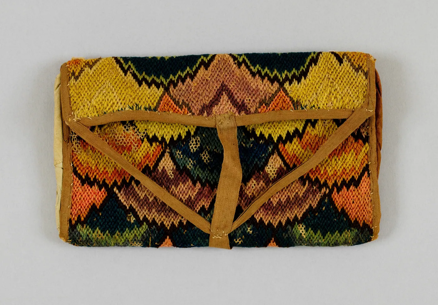 An eighteenth-century needlework pocketbook worked in a zigzag flame stitch of gold, rose, coral and deep green, closed with a tan fabric flap.
