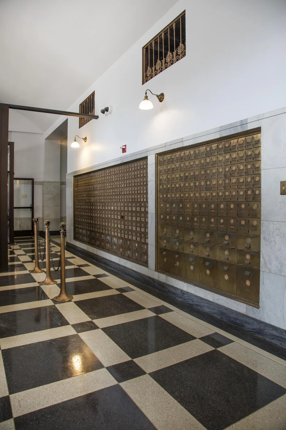 Two long banks of brass post office boxes set into a white marble wall above a black-and-white checkerboard terrazzo floor, lit by globe sconces.