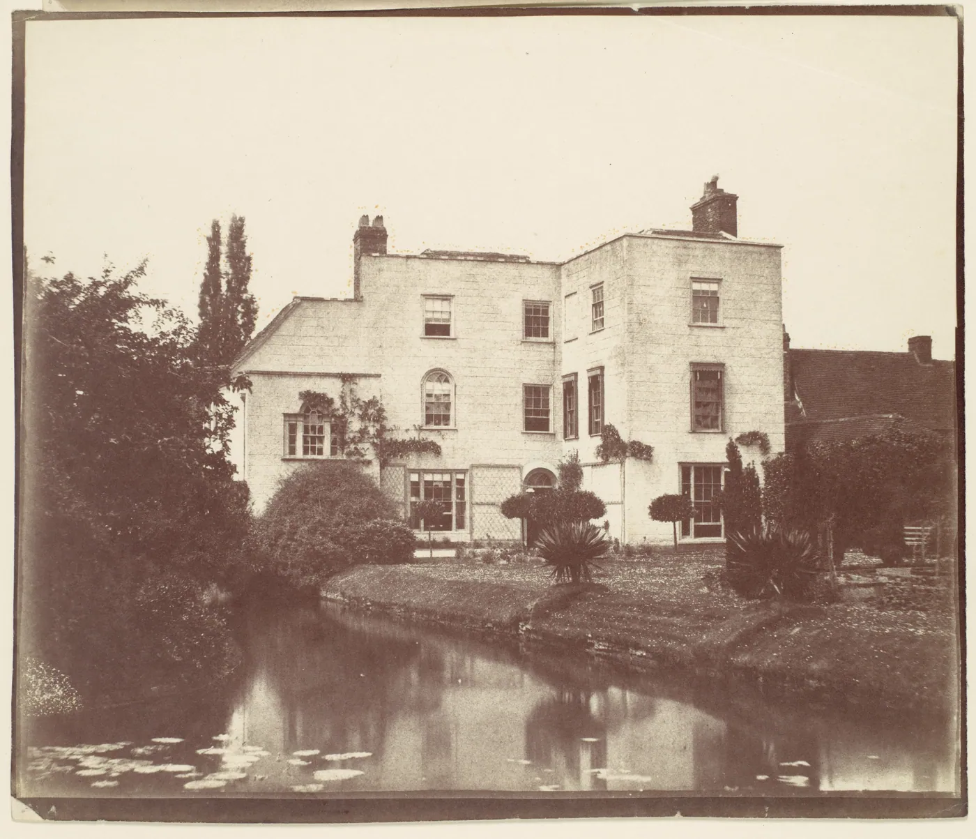 Sepia photograph of a three-storey pale stone house with tall chimneys, seen across a lawn and a still pond with lily pads, framed by shrubs and trees