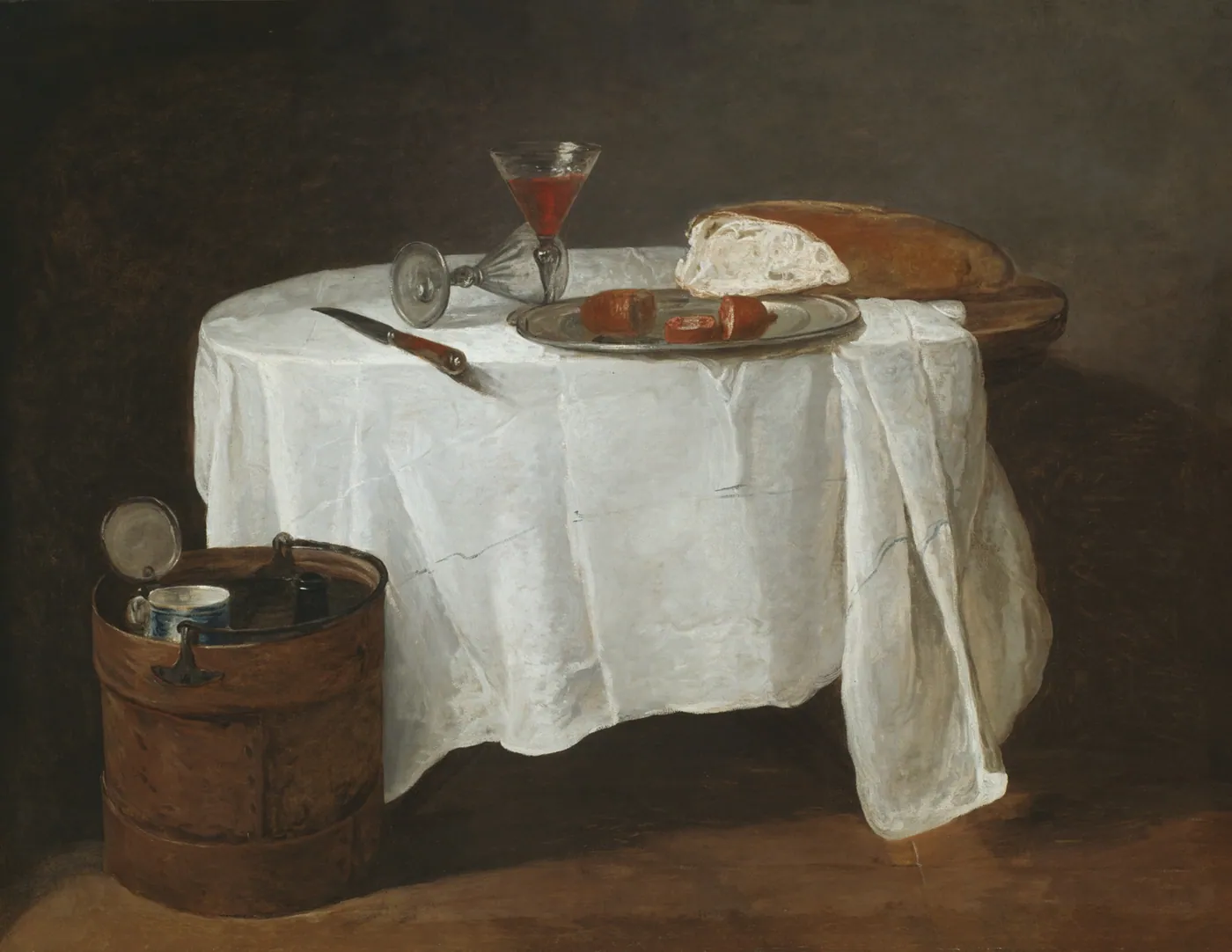 A small table draped in white linen holding a glass of red wine, bread and a plate, with a wooden wine cooler beside it.