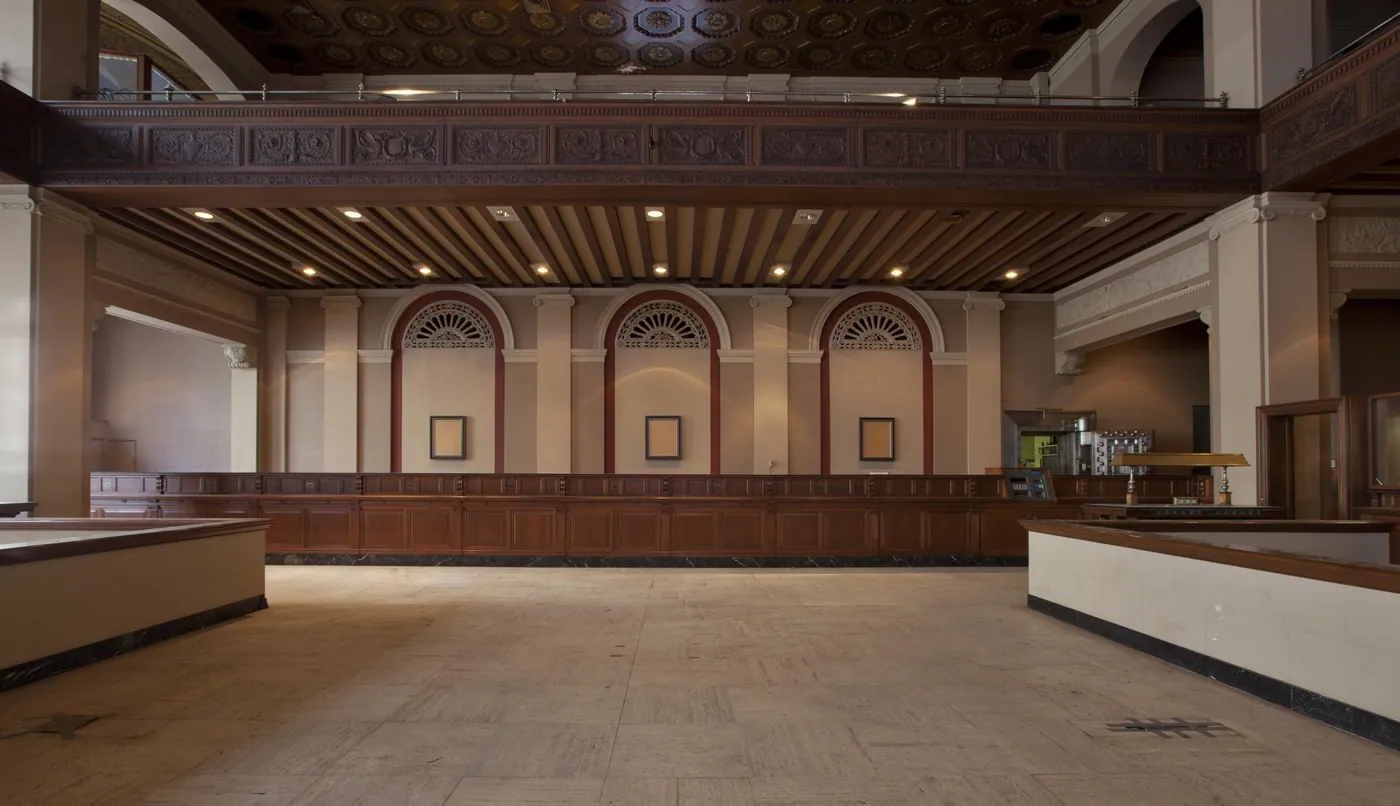 An empty 1920s banking hall with a long wooden teller counter, arched windows and a coffered, beamed ceiling.