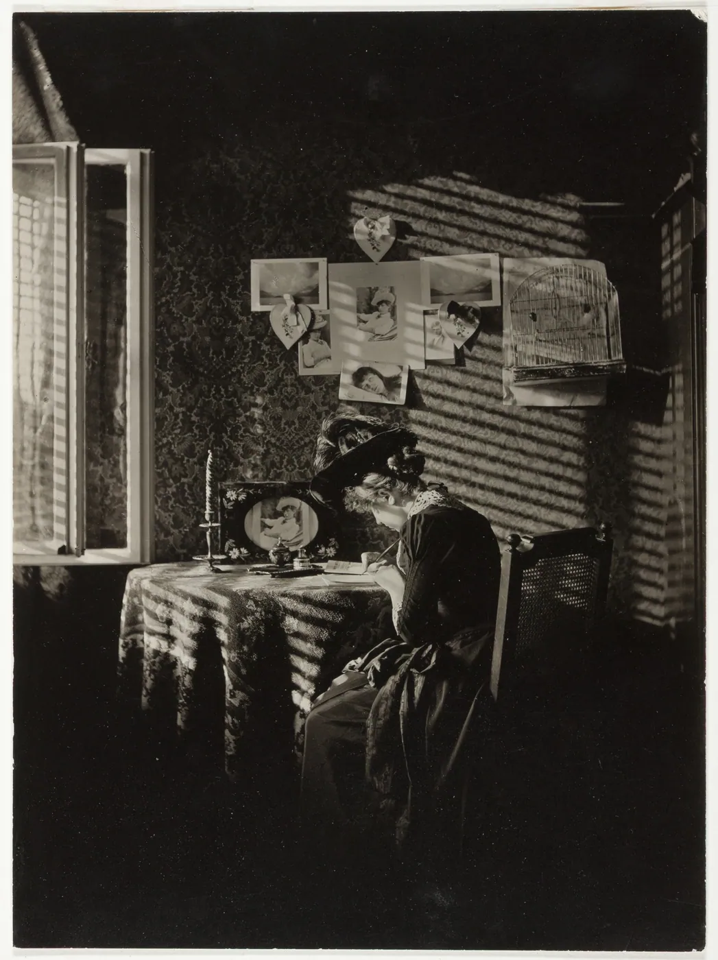 A woman writes at a lace-covered table by an open window, sunlight striping the wall where photographs and papers are pinned
