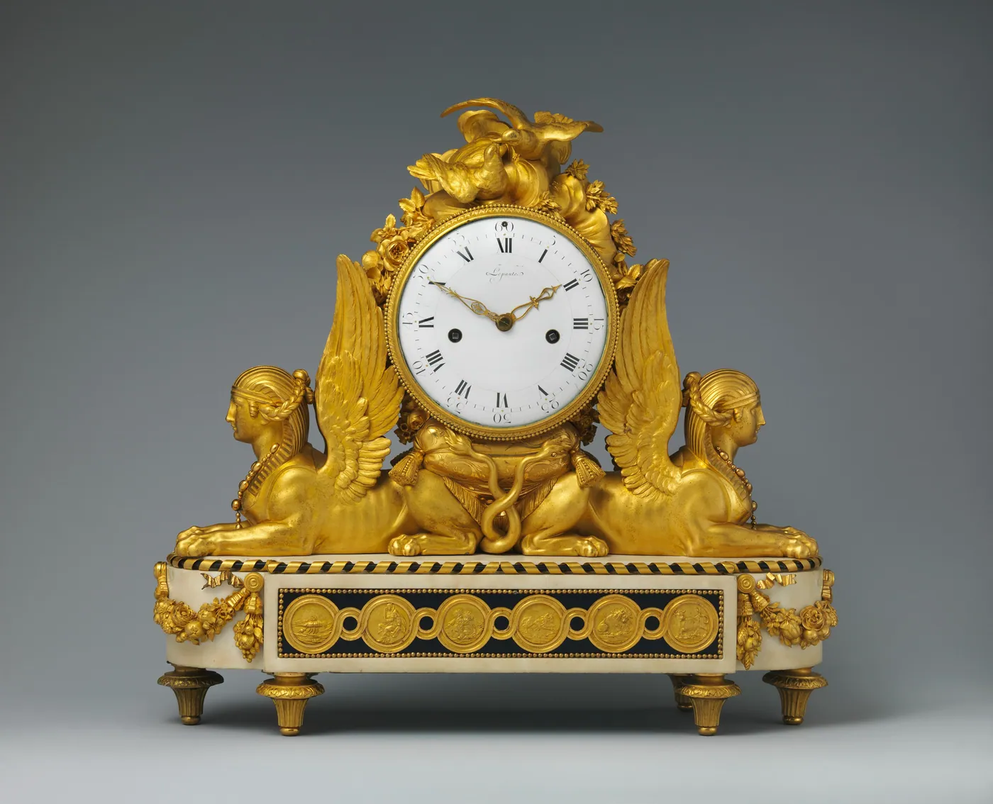 A gilt-bronze and white marble mantel clock with a white enamel dial flanked by two winged sphinxes, set against a grey background