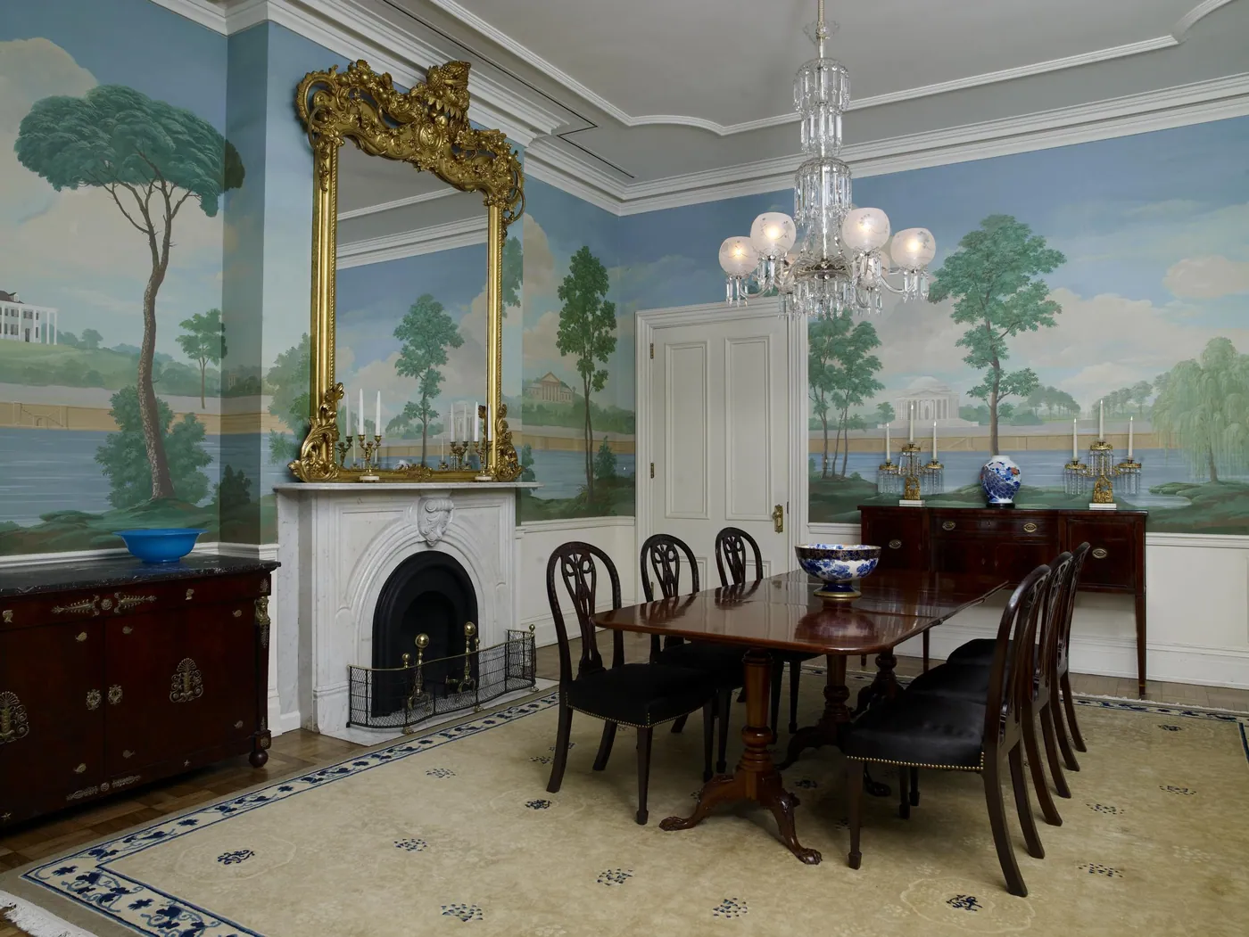 Formal dining room with a mahogany table and black-upholstered chairs, a gilt mirror over a marble fireplace, a crystal chandelier and scenic landscape wallpaper.