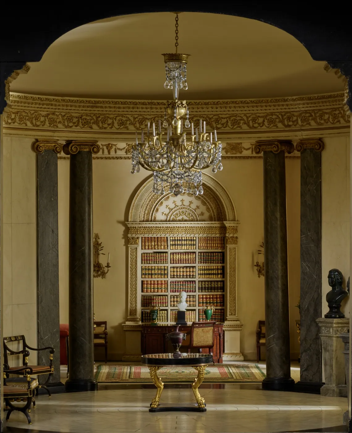 Miniature Regency rotunda with dark marble columns, a crystal chandelier, and a writing desk set before an arched bookcase of leather-bound volumes