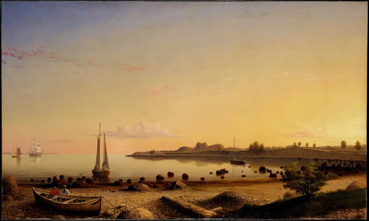 A still, golden harbor at sunset with sailboats at anchor, a rocky point across the water, and a small rowboat drawn up on the stony shore in the foreground.