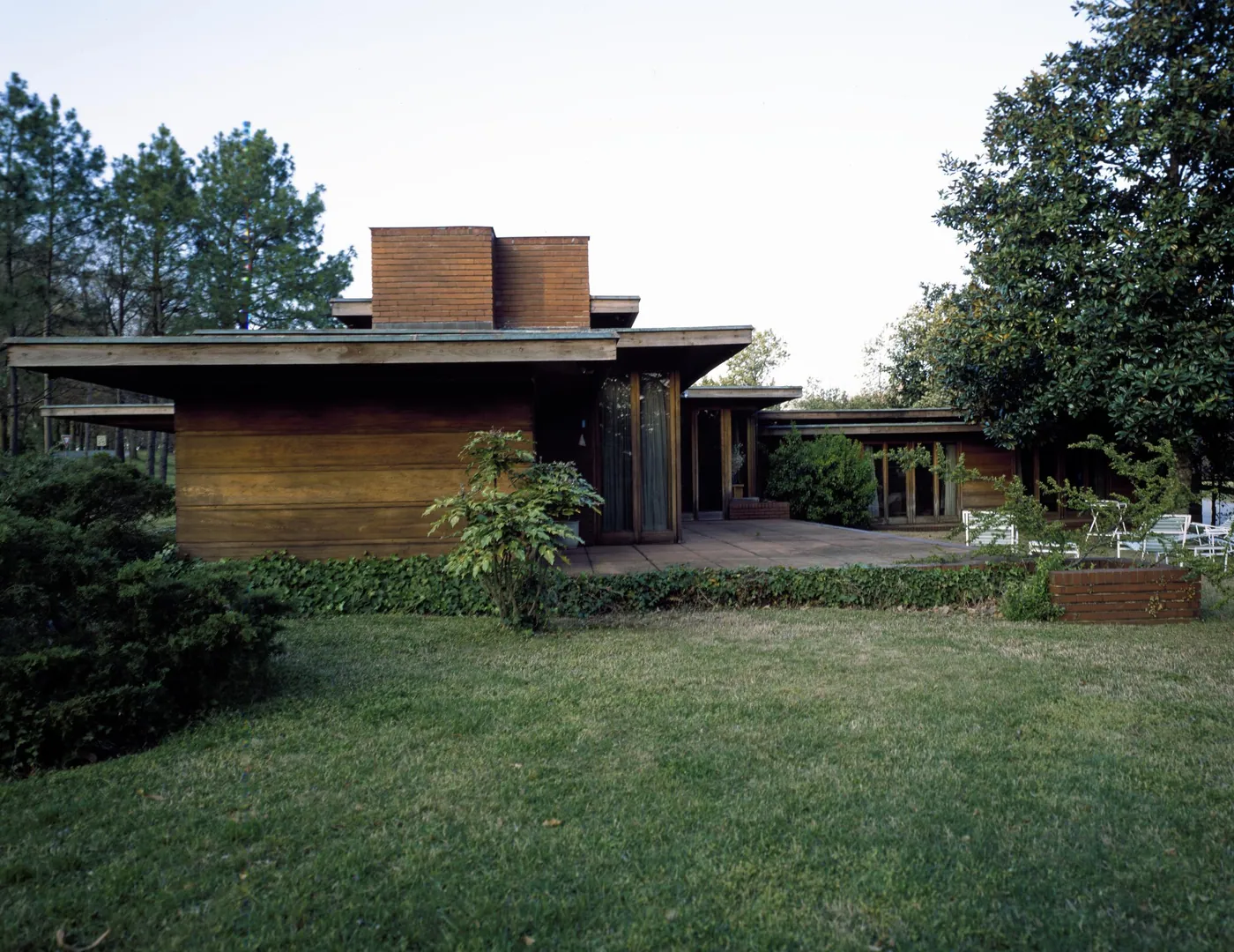 Low cypress-and-brick Usonian house with a broad flat roof and brick chimney, set behind a lawn with a terrace of white garden chairs