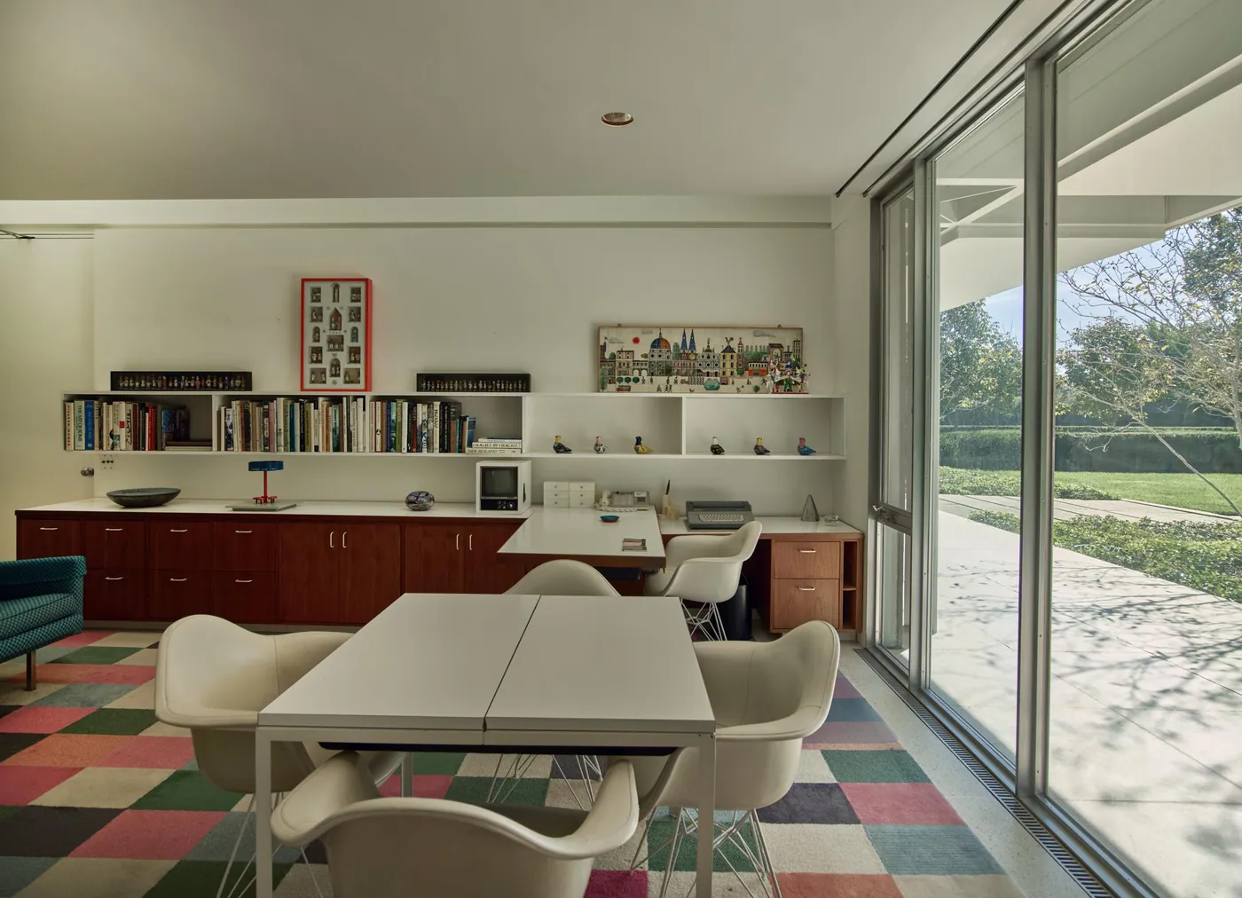 White shell armchairs around a white table on a patchwork carpet, with walnut cabinets, bookshelves and a glass wall onto the lawn