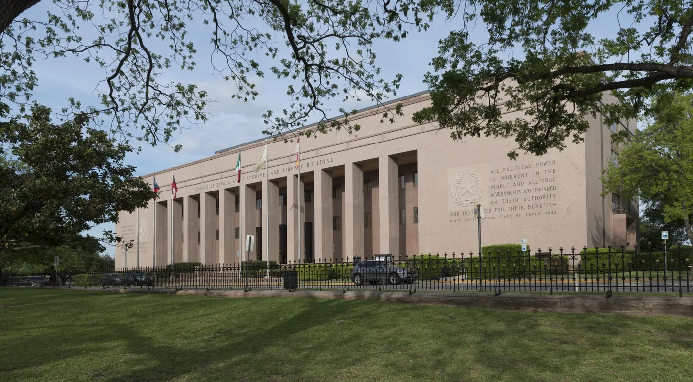 A long pink-granite archives building with tall square columns and a carved inscription, framed by oak branches above a lawn.