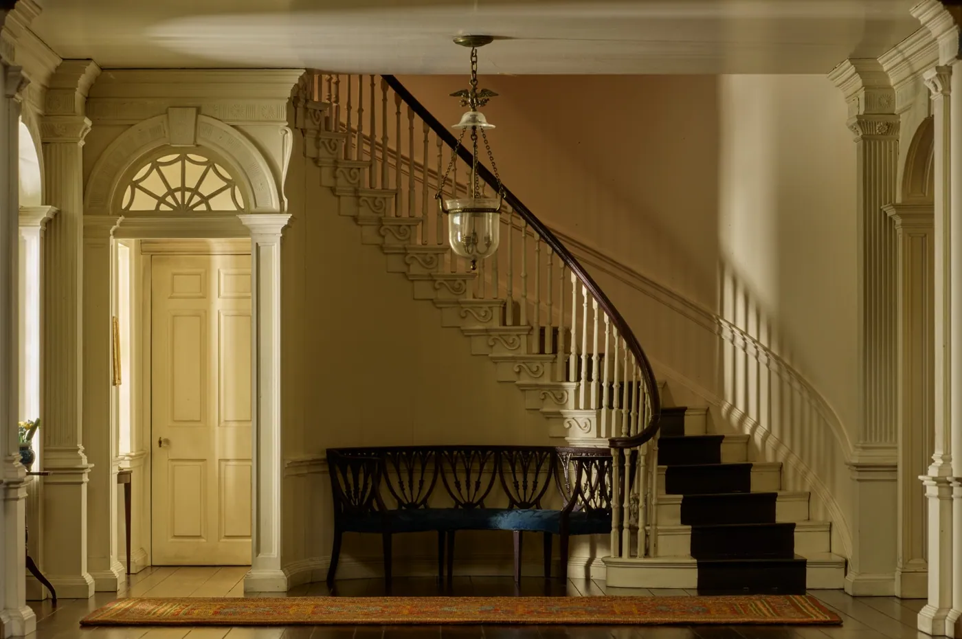 Miniature entrance hall with a fanlit panelled door, a curving staircase with a dark handrail, a lantern hanging overhead, a bench and a long runner rug