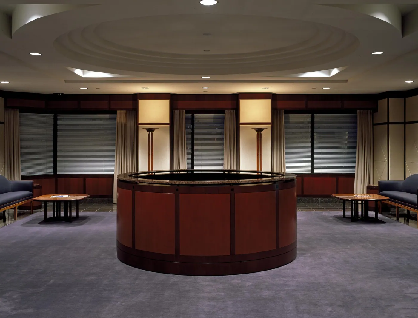 Quiet office reception area with a round mahogany desk at center, low wooden tables and upholstered benches, and tall windows with blinds and drapes.
