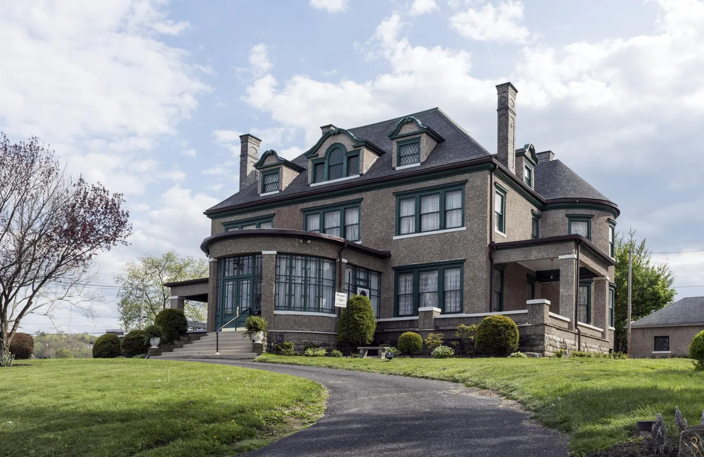 A stately stone clubhouse with dormers, green-trimmed windows and a curved glass porch, set on a lawn under a soft blue sky