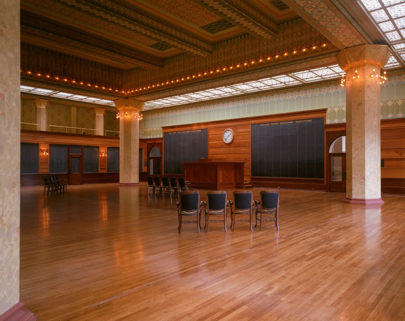 Empty wood-floored trading hall with stencilled columns, a skylit ceiling ringed with lights, a wall clock above dark quotation boards, and a few chairs.