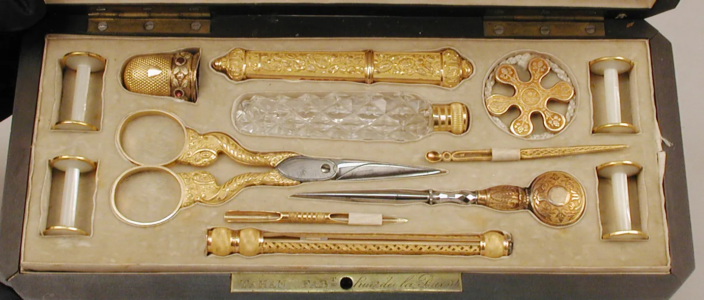 An open fitted case lined in cream velvet, holding gold-mounted scissors, a thimble, needle cases, a crystal flask and thread spools, each in its own slot.