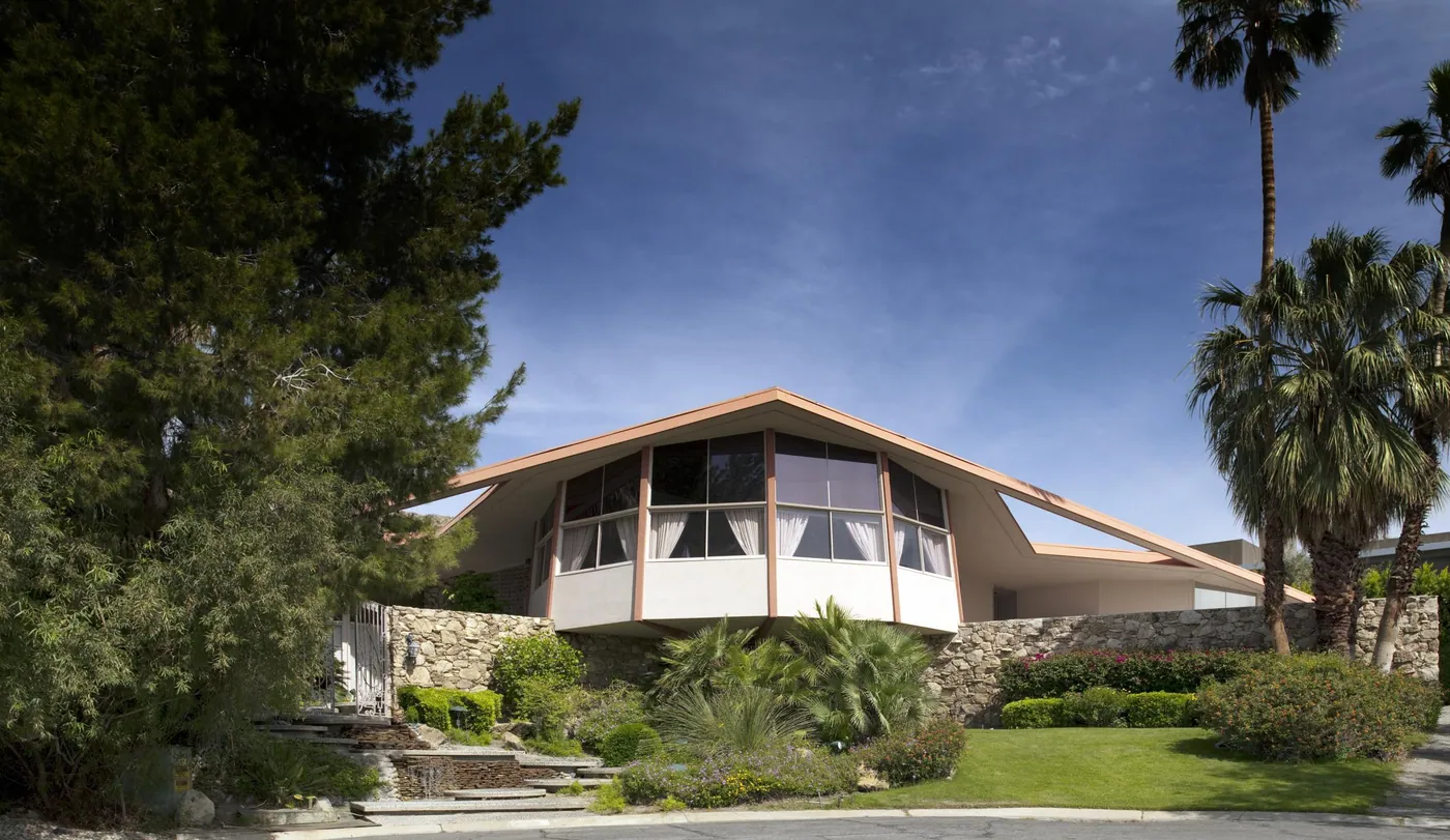 Mid-century house with a sweeping angled roof over a curved bank of windows, set on a stone base among palms and garden plantings