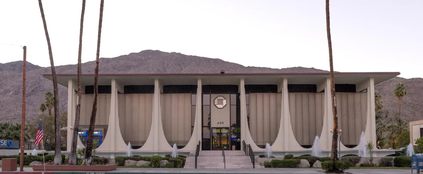 Low white modernist bank with flared parabolic columns, fountains and palms, set against the San Jacinto mountains at dusk