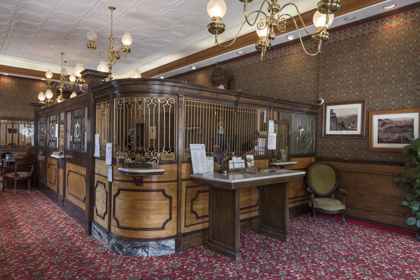 A wood-panelled bank interior with brass-grilled teller windows, a marble-topped writing table, chandeliers and a red patterned carpet