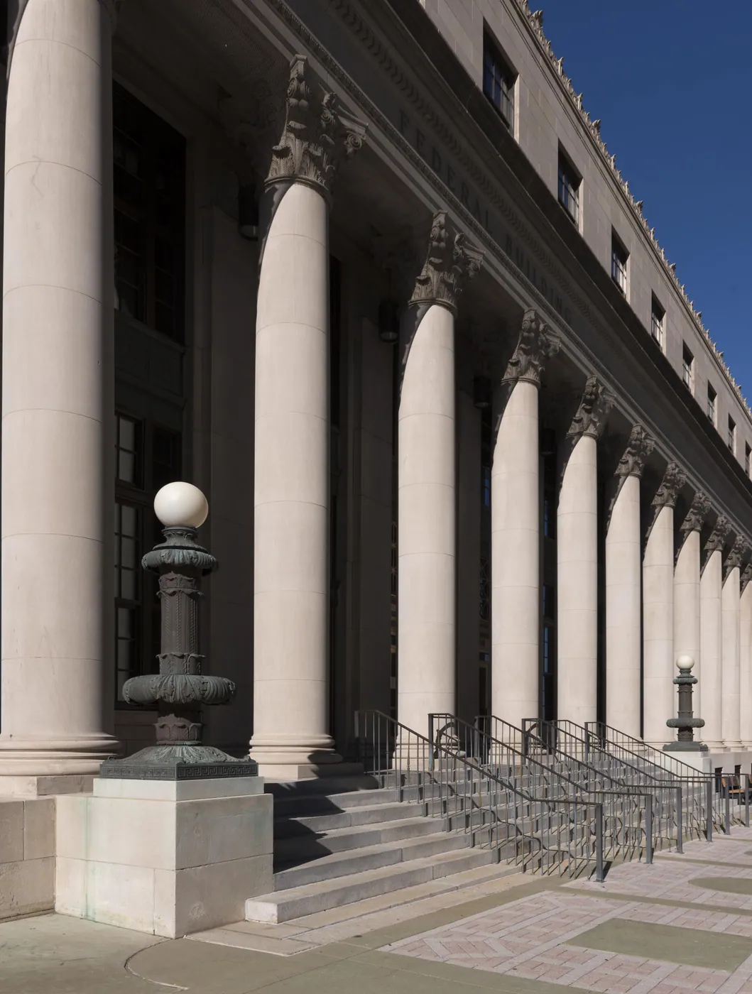 A long colonnade of tall limestone columns with carved capitals rises beside broad entrance steps and bronze lamp standards under a deep blue sky.
