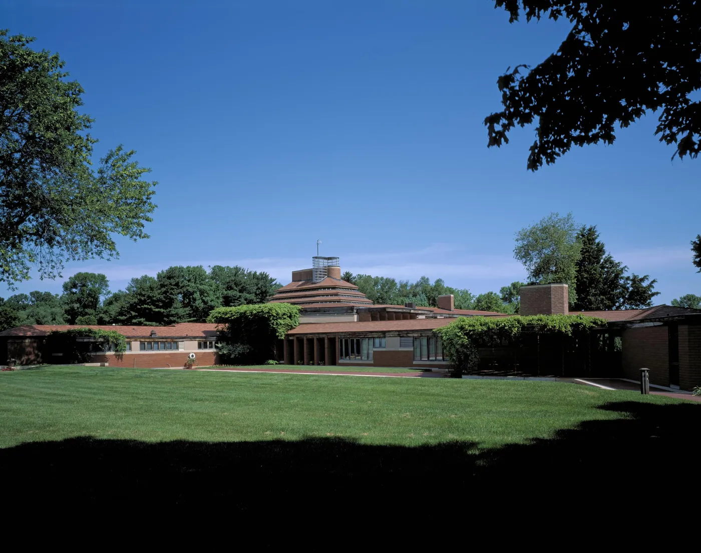 Long, low brick house with a stepped central chimney tower, set behind a wide green lawn under a deep blue sky and framing trees
