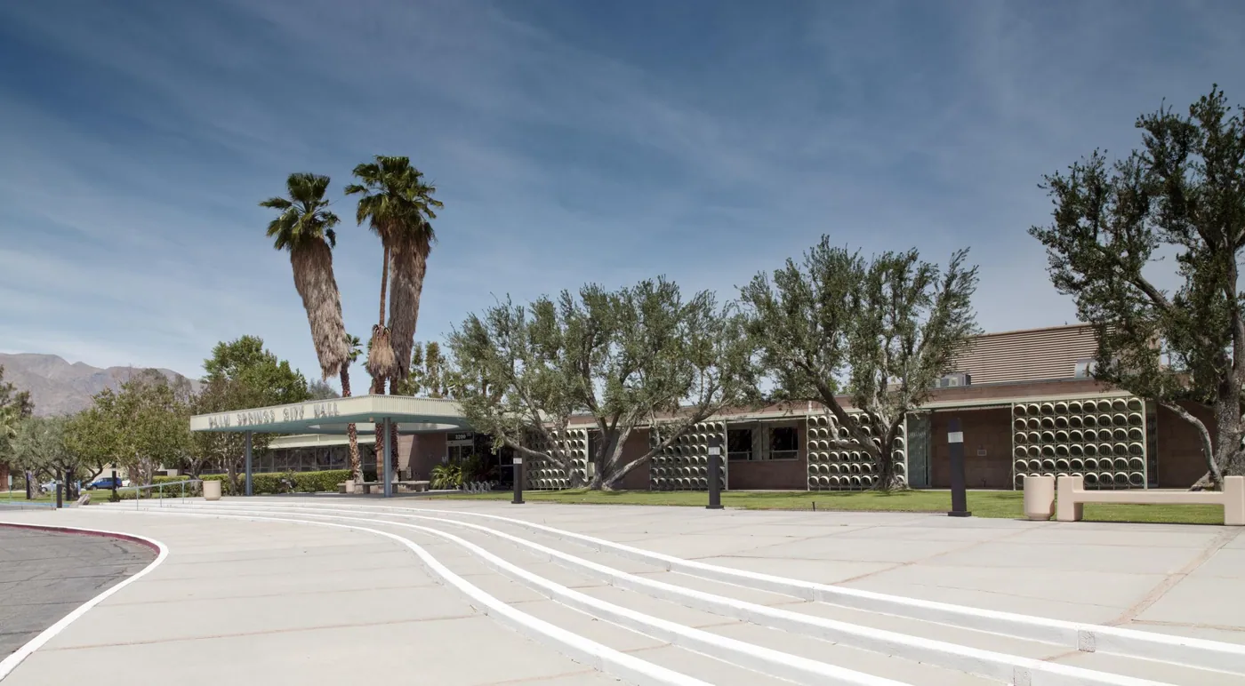 Low modernist civic building with patterned block screens and a flat entrance canopy, olive trees and tall palms, and a wide curving pale plaza