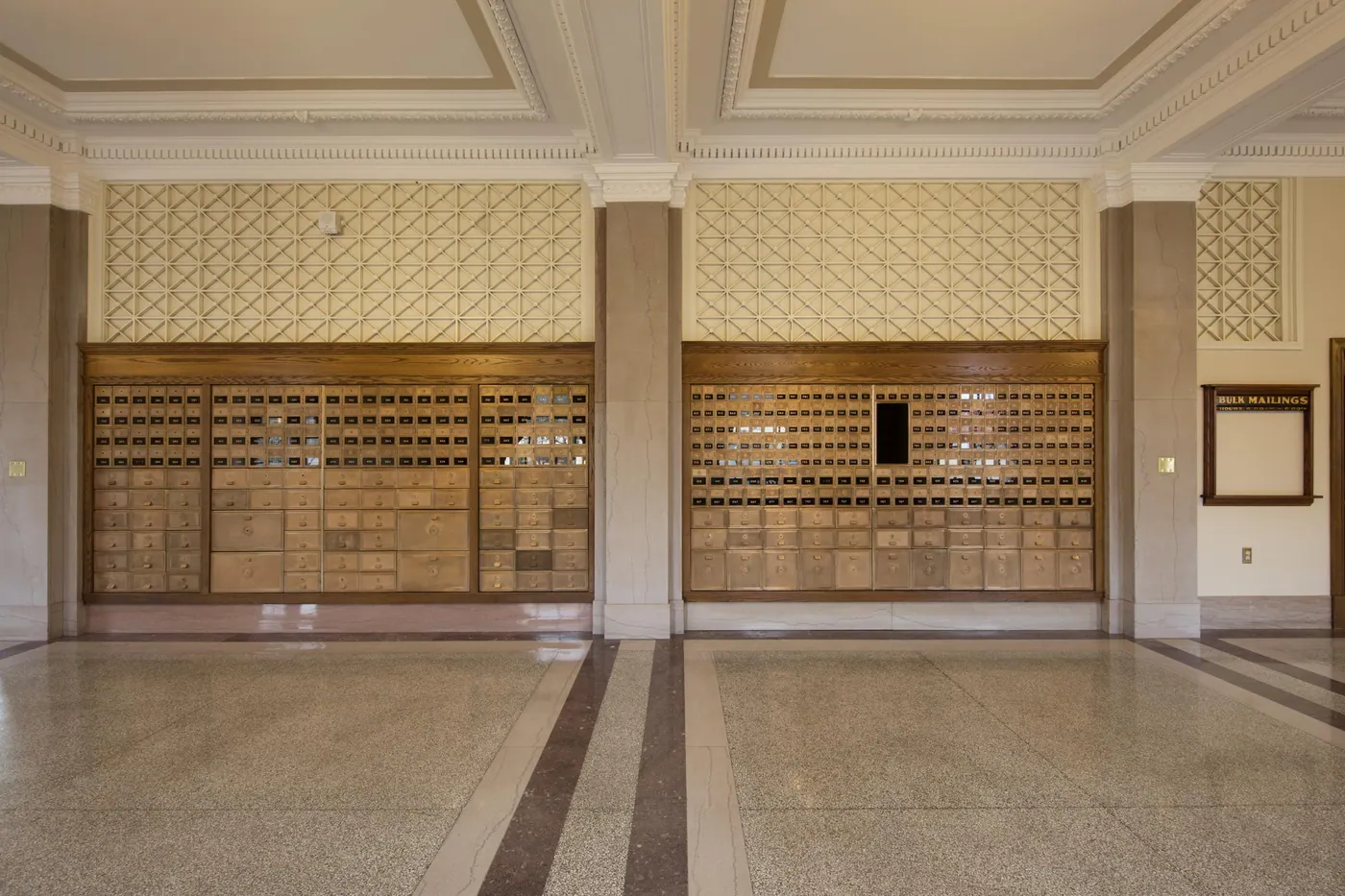 Two symmetrical banks of brass and wood post office boxes set between marble pilasters in a quiet lobby with a polished terrazzo floor