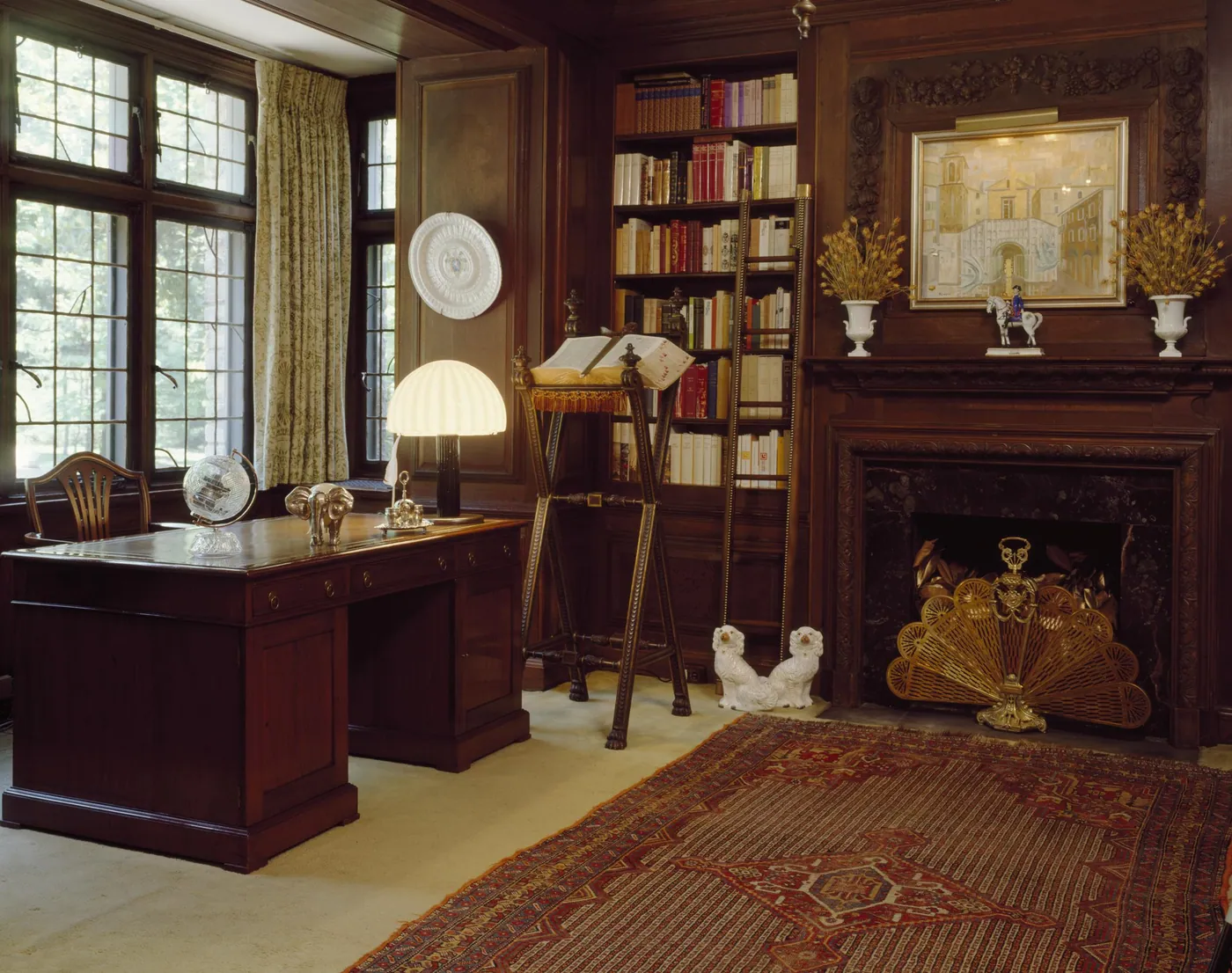 Panelled study with a partners' desk and dome lamp by leaded windows, an open book on a lectern, bookshelves and a carved fireplace