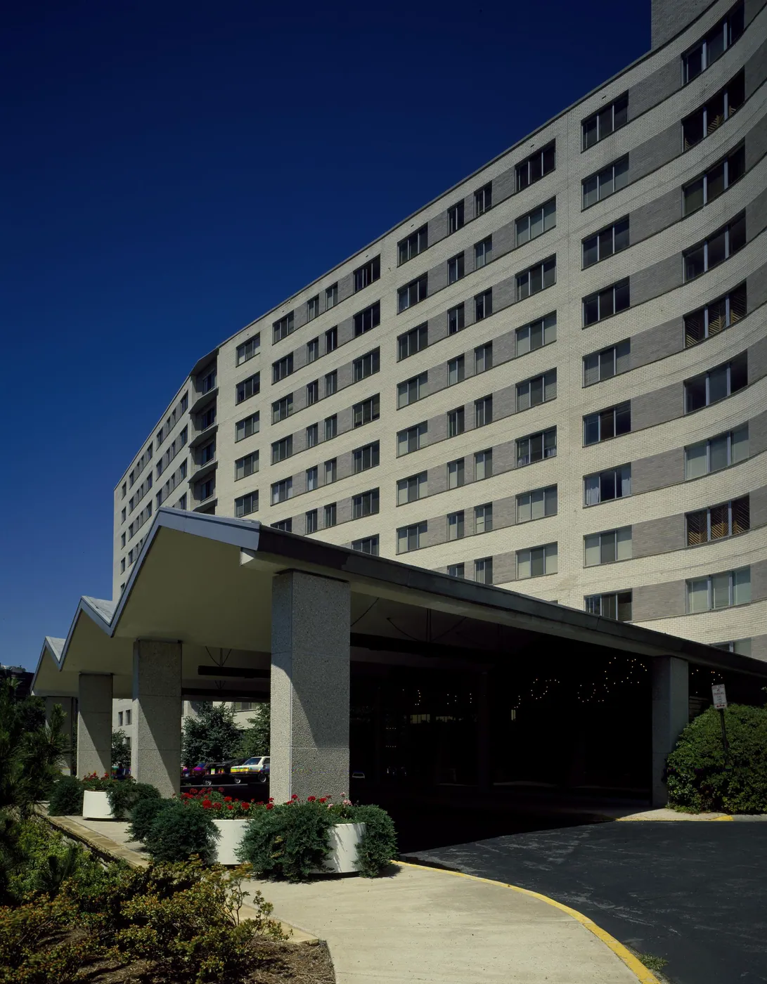 A curving modernist apartment tower of pale brick against a deep blue sky, with a zigzag-roofed concrete porte-cochère and planters of red flowers at the entrance.