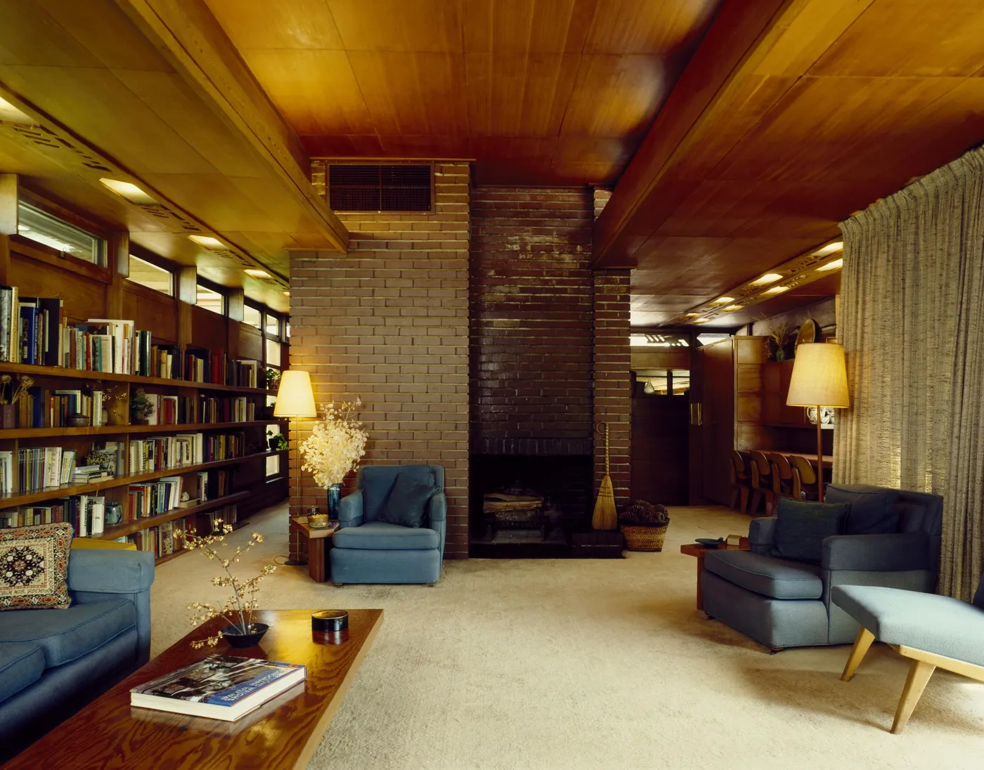 Warm wood-ceilinged living room with a brick fireplace, long built-in bookshelves, blue armchairs and sofa, lamps and a low wooden coffee table