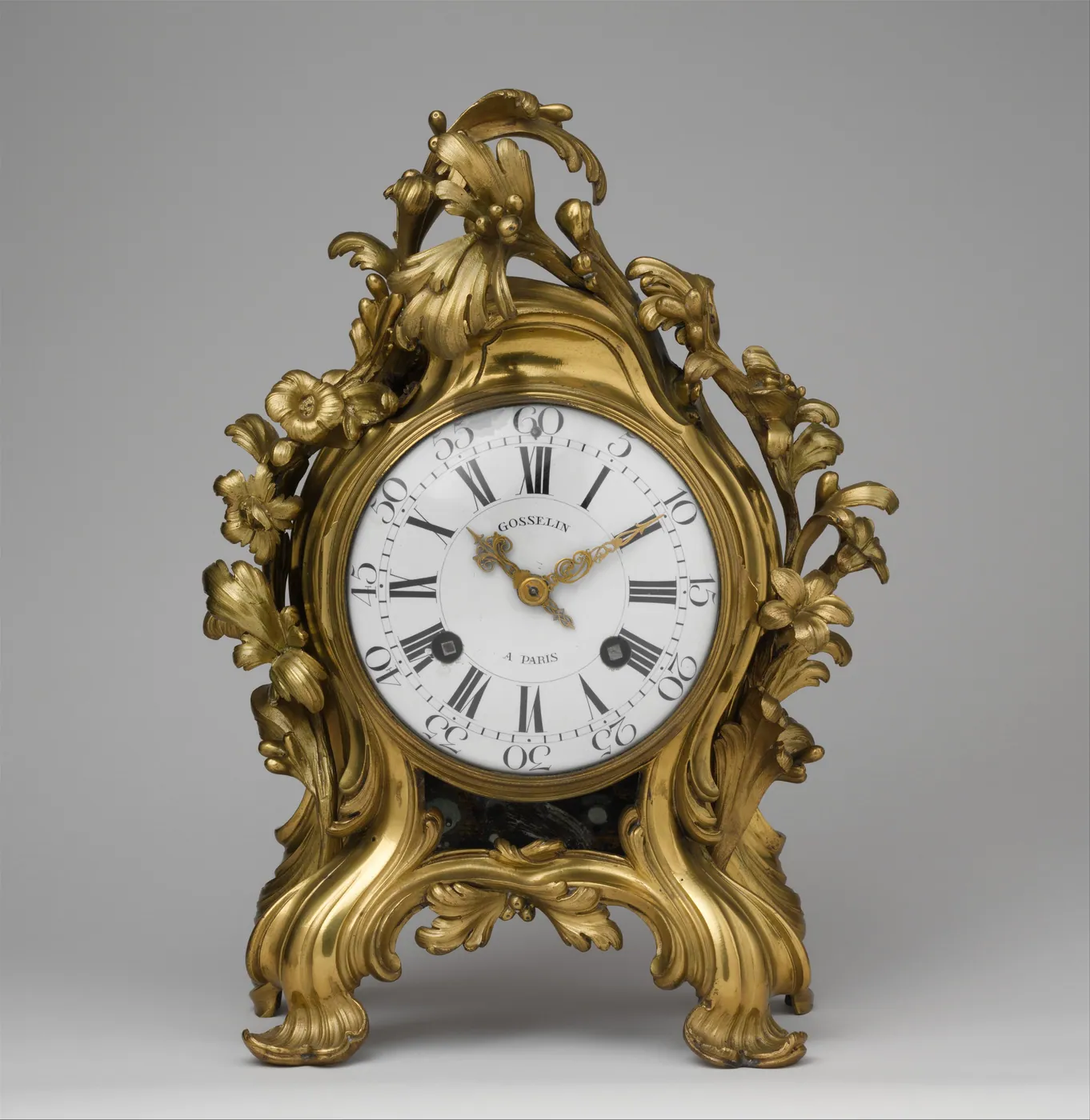 Gilt-bronze Rococo mantel clock with a white enamel dial and Roman numerals, framed by cast leaves and flowers, on a plain grey background
