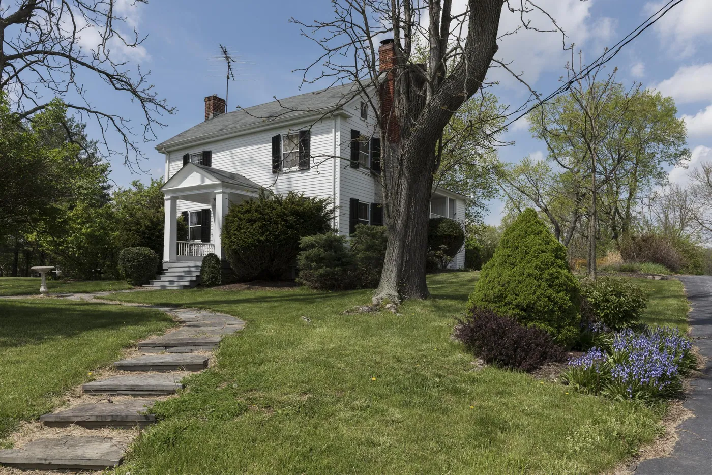 A white two-storey farmhouse with black shutters and a columned porch, reached by a stone path across a spring lawn with shrubs and bluebells