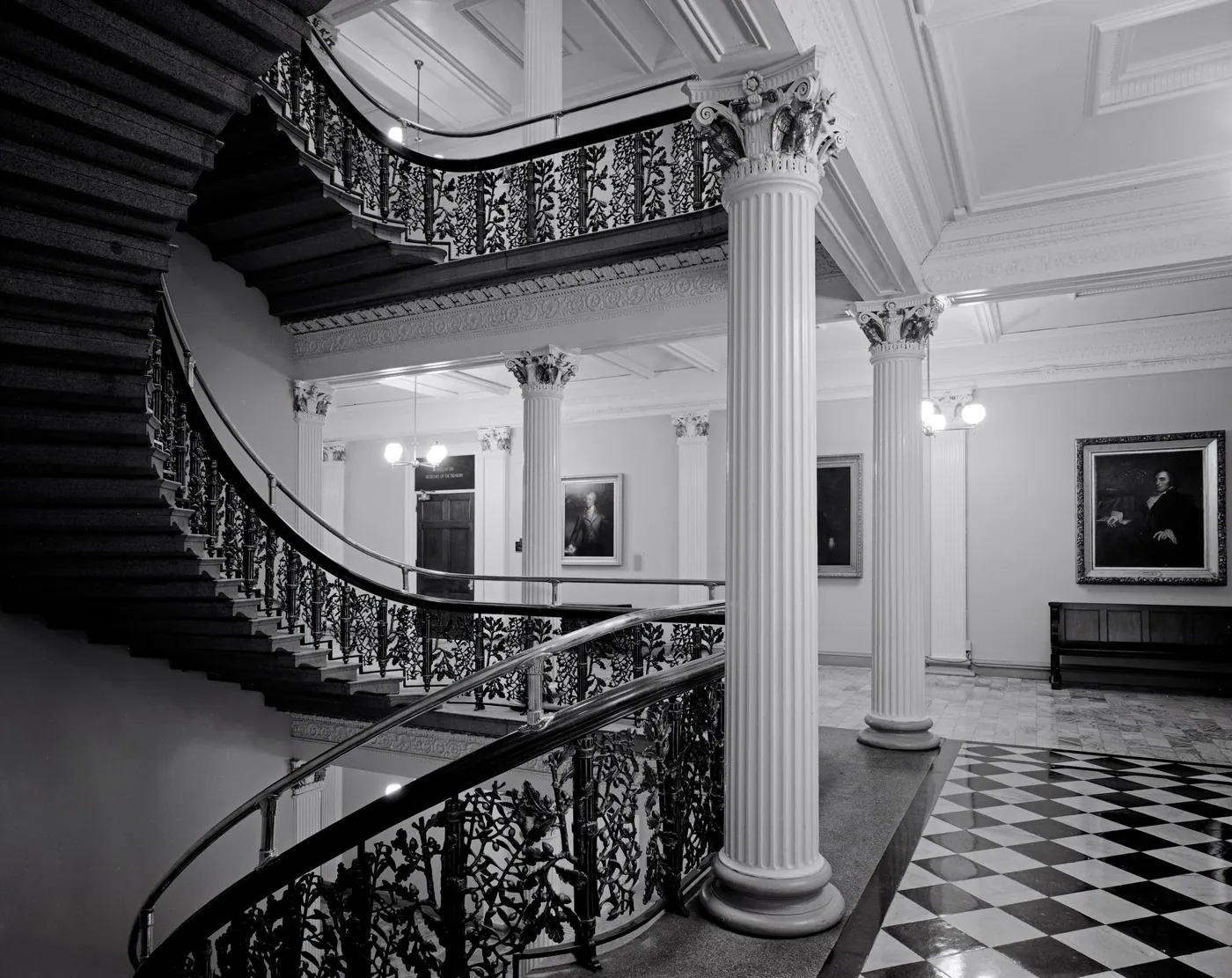 A black-and-white view of a curving staircase with ornate iron railings, fluted columns and a checkerboard marble floor inside the Treasury building.