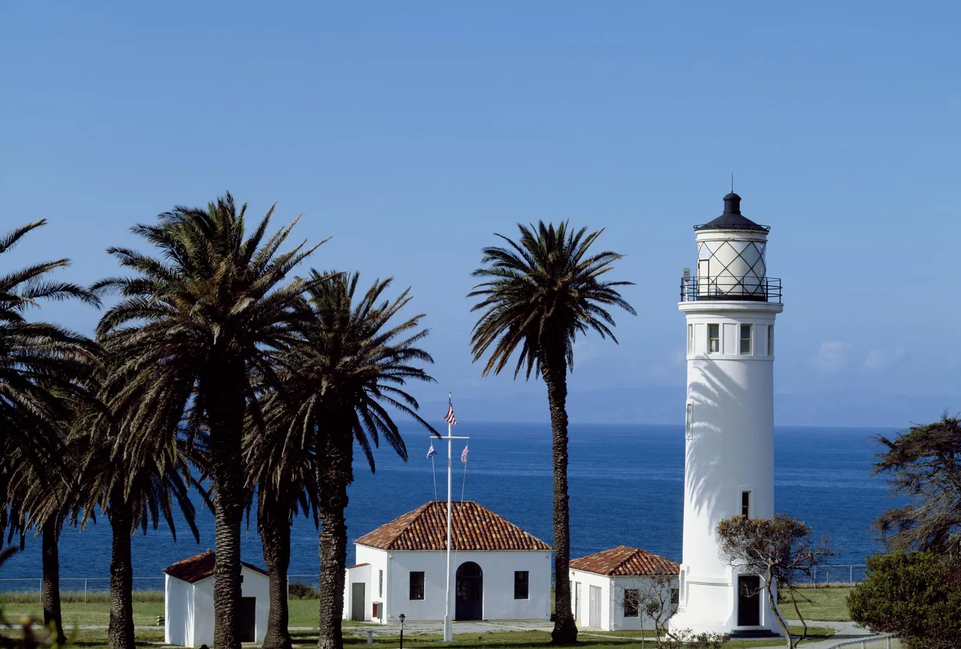 A white lighthouse tower and red-roofed keepers' buildings among tall palms on a bluff above a deep blue Pacific.