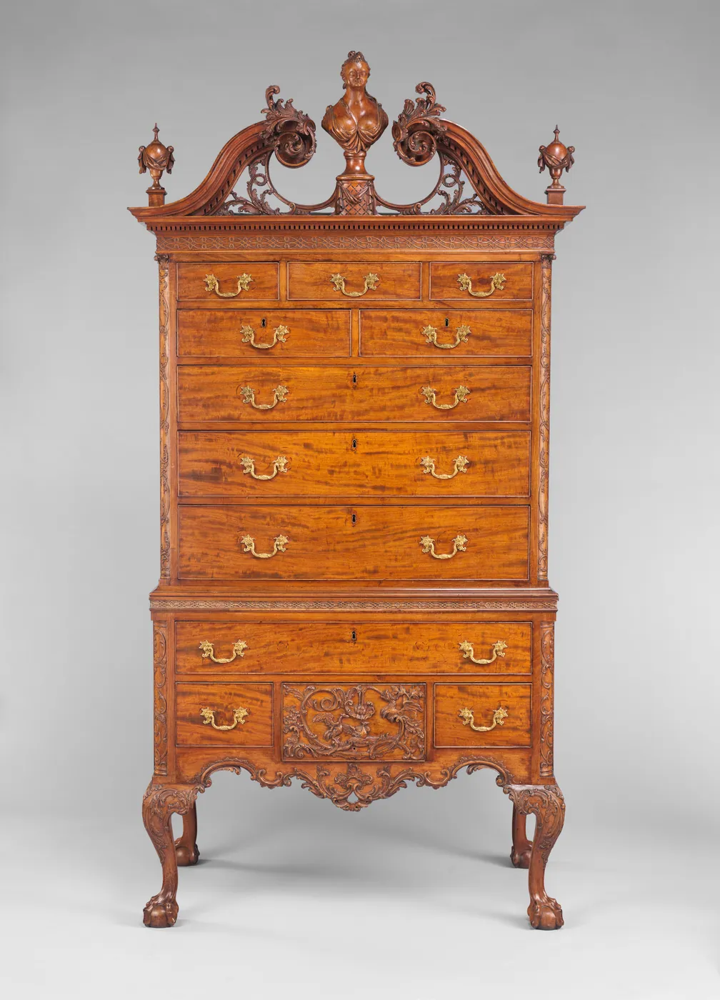 A tall mahogany high chest with a carved scrolled pediment, central bust finial, brass drawer pulls and claw-and-ball feet, shown on a grey backdrop