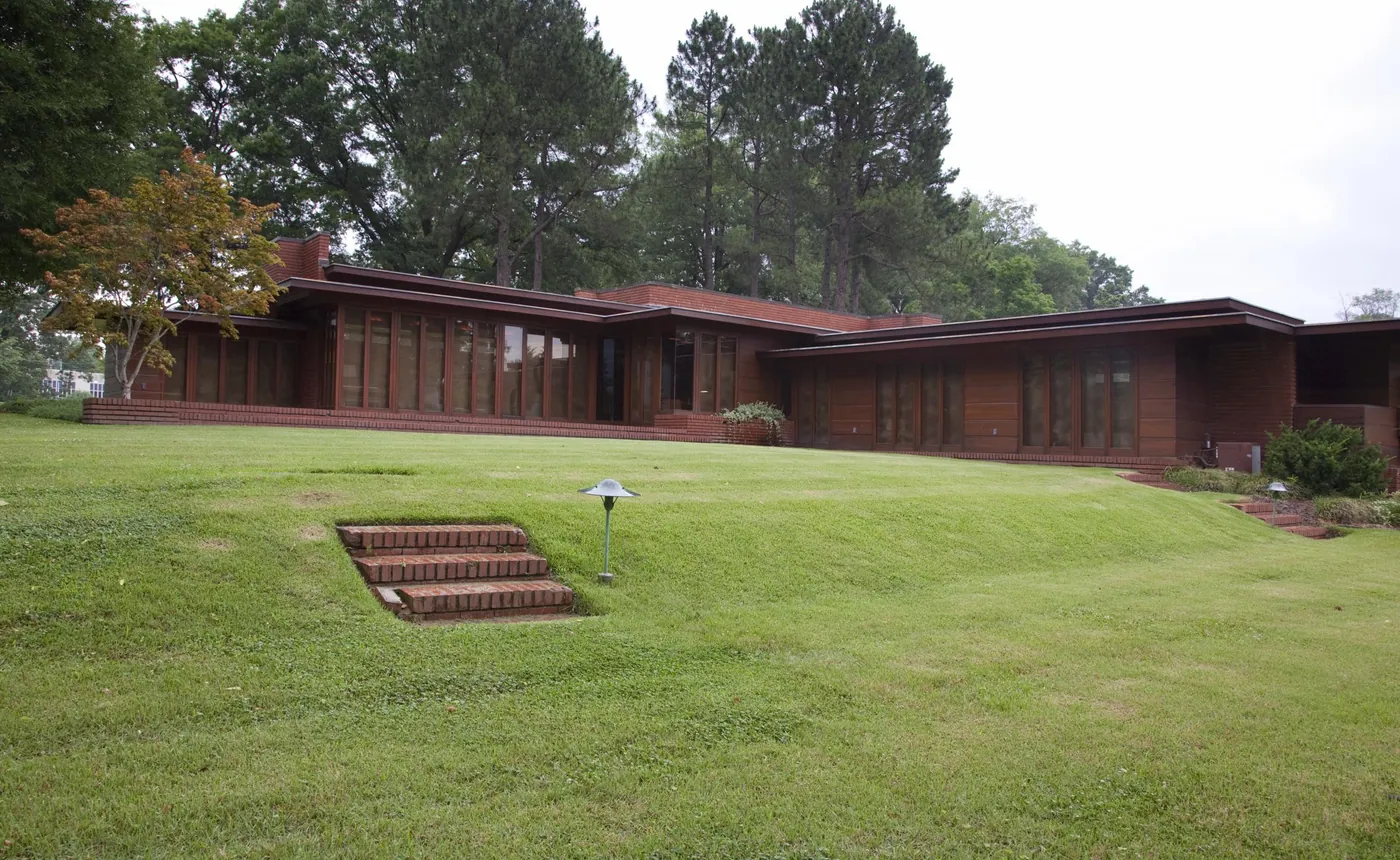 Frank Lloyd Wright's long, low brick-and-cypress Rosenbaum House behind a sloping green lawn with brick steps, tall pines and a red-leafed tree around it