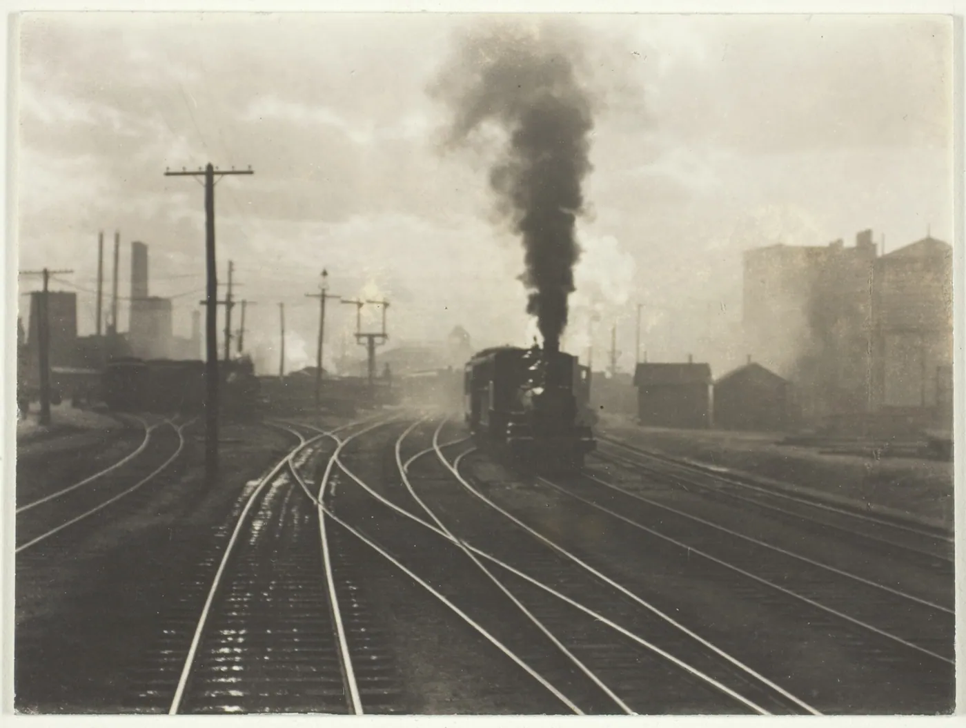 A steam locomotive trails black smoke through a hazy rail yard where many sets of gleaming tracks curve, cross and branch apart