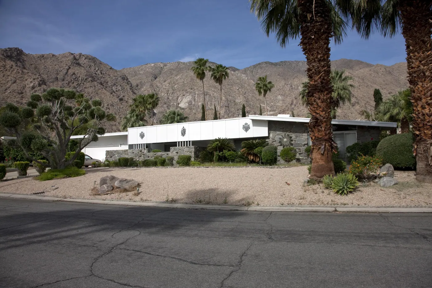A low white mid-century modern house with decorative fascia medallions and stone walls, framed by palms and desert gardens beneath the San Jacinto mountains