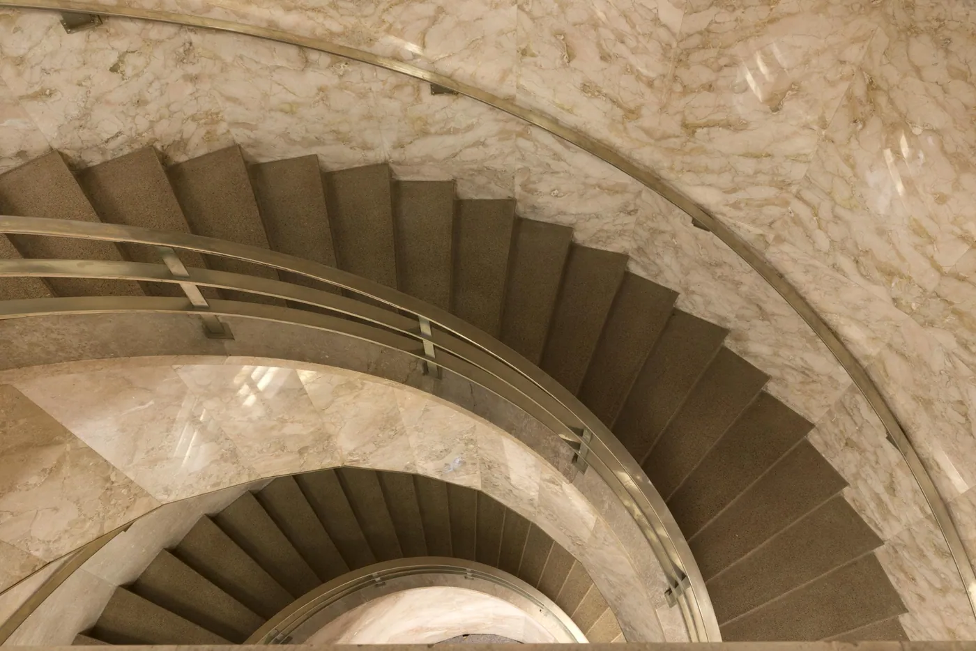 View down a curving staircase with a brass handrail, spiralling between walls of pale, gold-veined polished marble