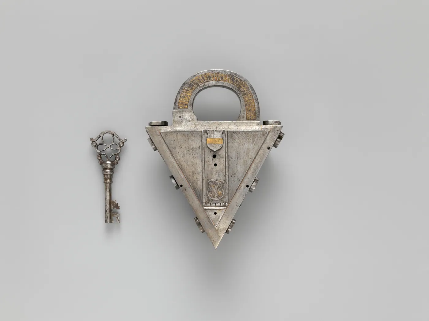 Triangular steel padlock with a gilded, engraved shackle, shown beside its ornate pierced key on a plain grey background