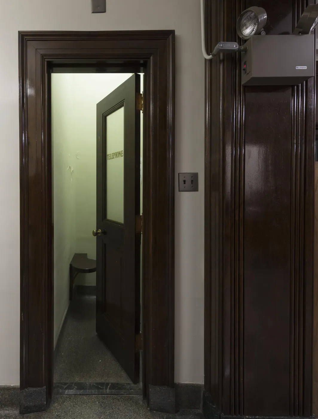 Dark-stained wooden doorway opening onto a small telephone booth with a half-open glazed door and a built-in seat