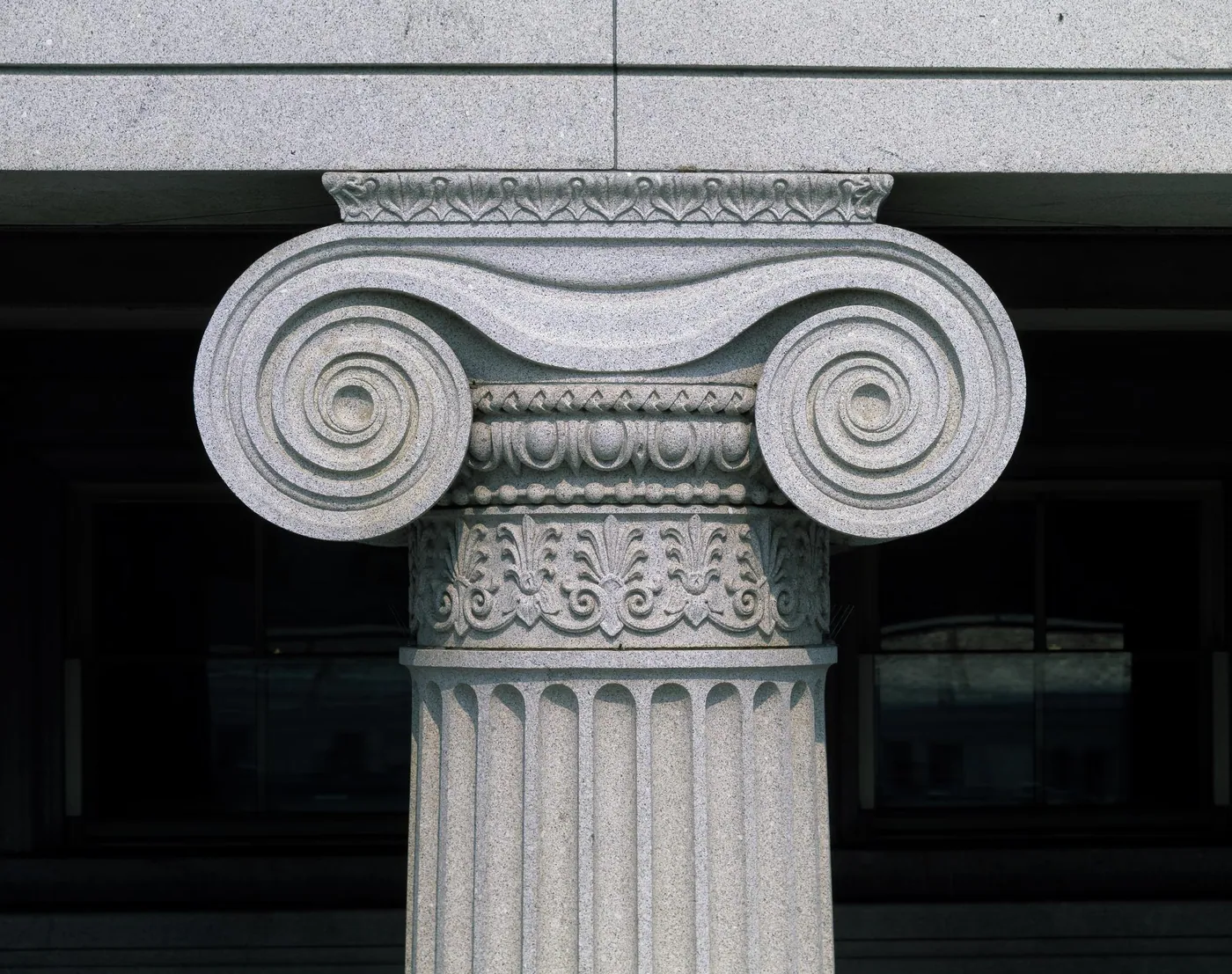 Close view of a carved granite Ionic column capital with deep spiral volutes above a fluted shaft, set against a shadowed facade