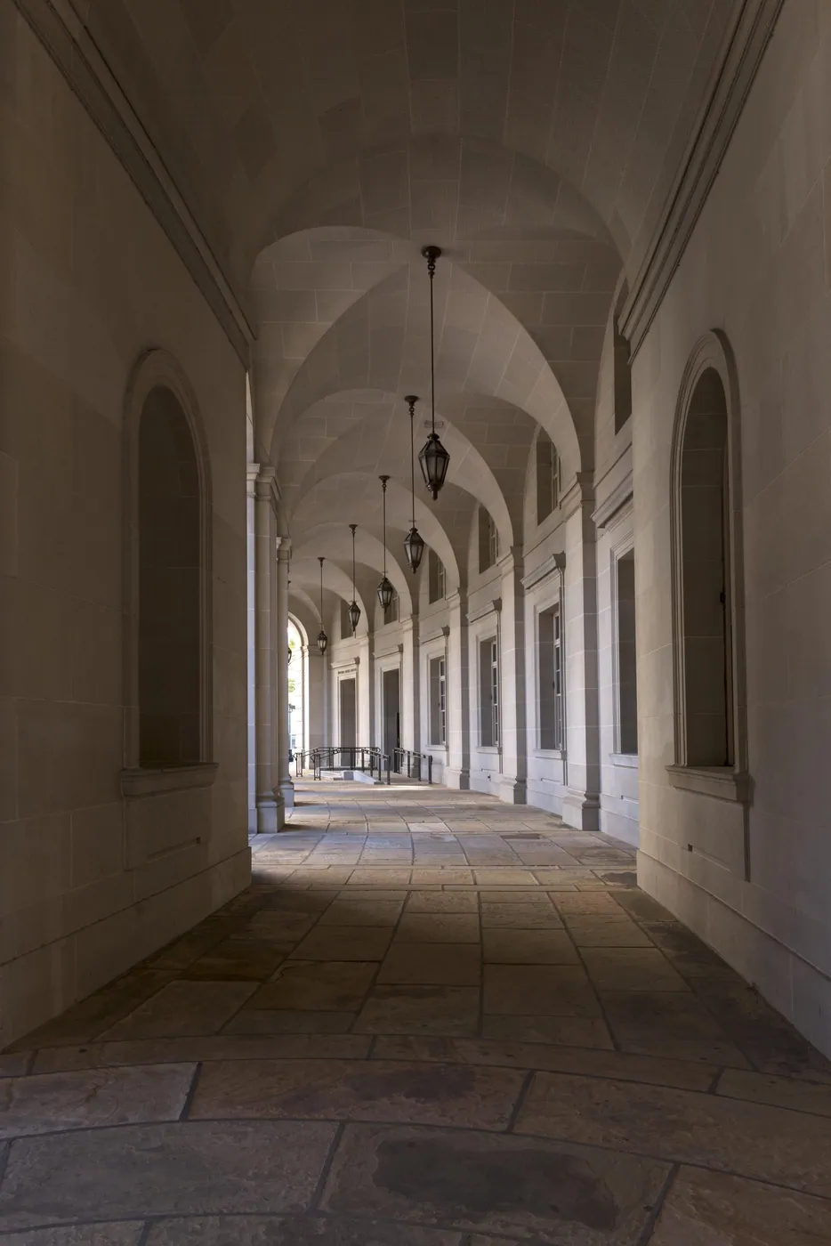 A long vaulted stone arcade with hanging lanterns and tall arched windows, receding in soft light toward a distant opening