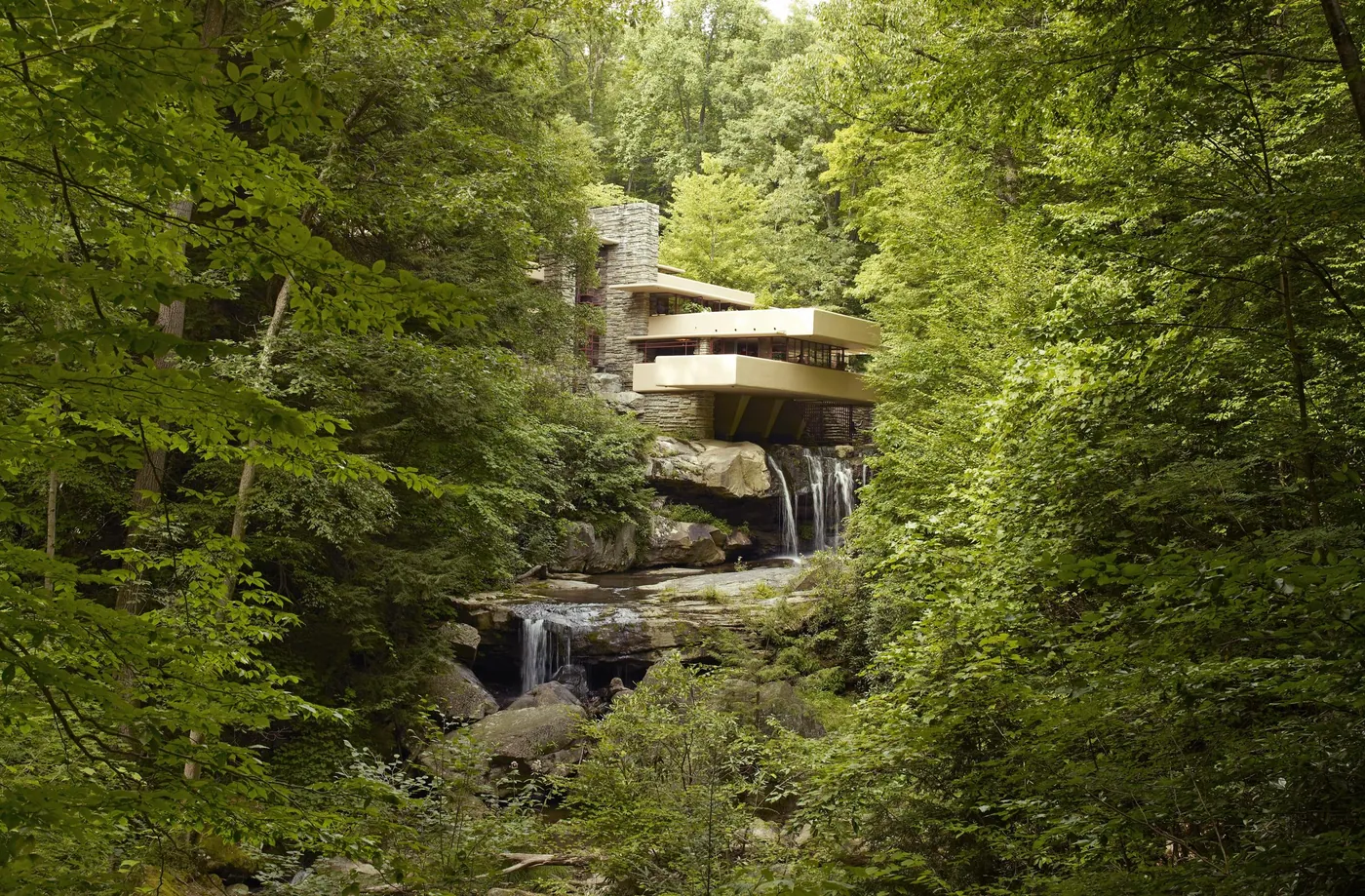 Frank Lloyd Wright's Fallingwater, its cream cantilevered terraces and stone chimney rising over a waterfall, seen through dense summer woodland