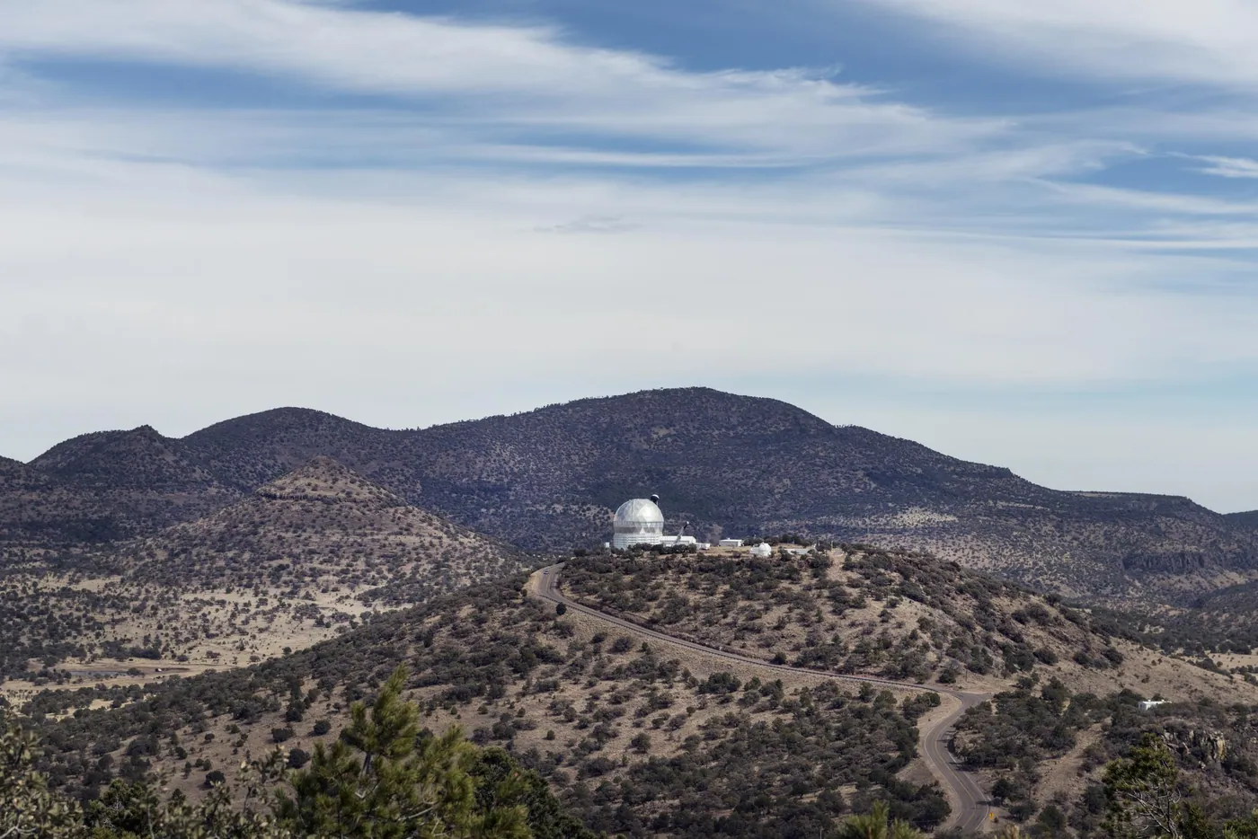 A white observatory dome on a hilltop in the dry Davis Mountains of West Texas, reached by a winding road beneath a wide sky of streaked cloud