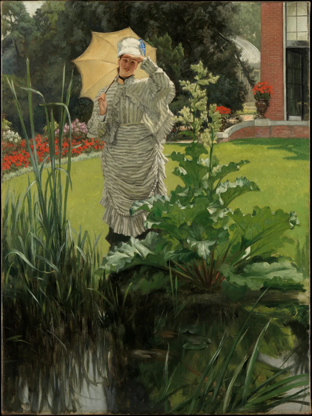 A woman in a striped summer dress holding a pale parasol walks across a sunny lawn beside a pond, with red flower beds and a brick house behind.