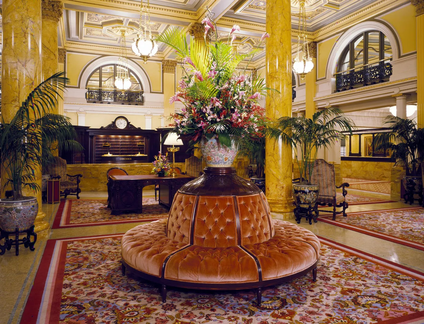 Grand hotel lobby with marble columns, a round tufted velvet banquette under a tall flower arrangement, palms, and a reception desk behind