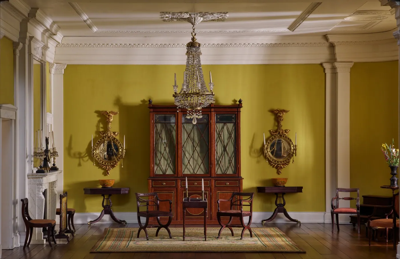 Miniature period drawing room with olive walls, a crystal chandelier, a glass-fronted secretary bookcase, gilt mirrors, and mahogany chairs.