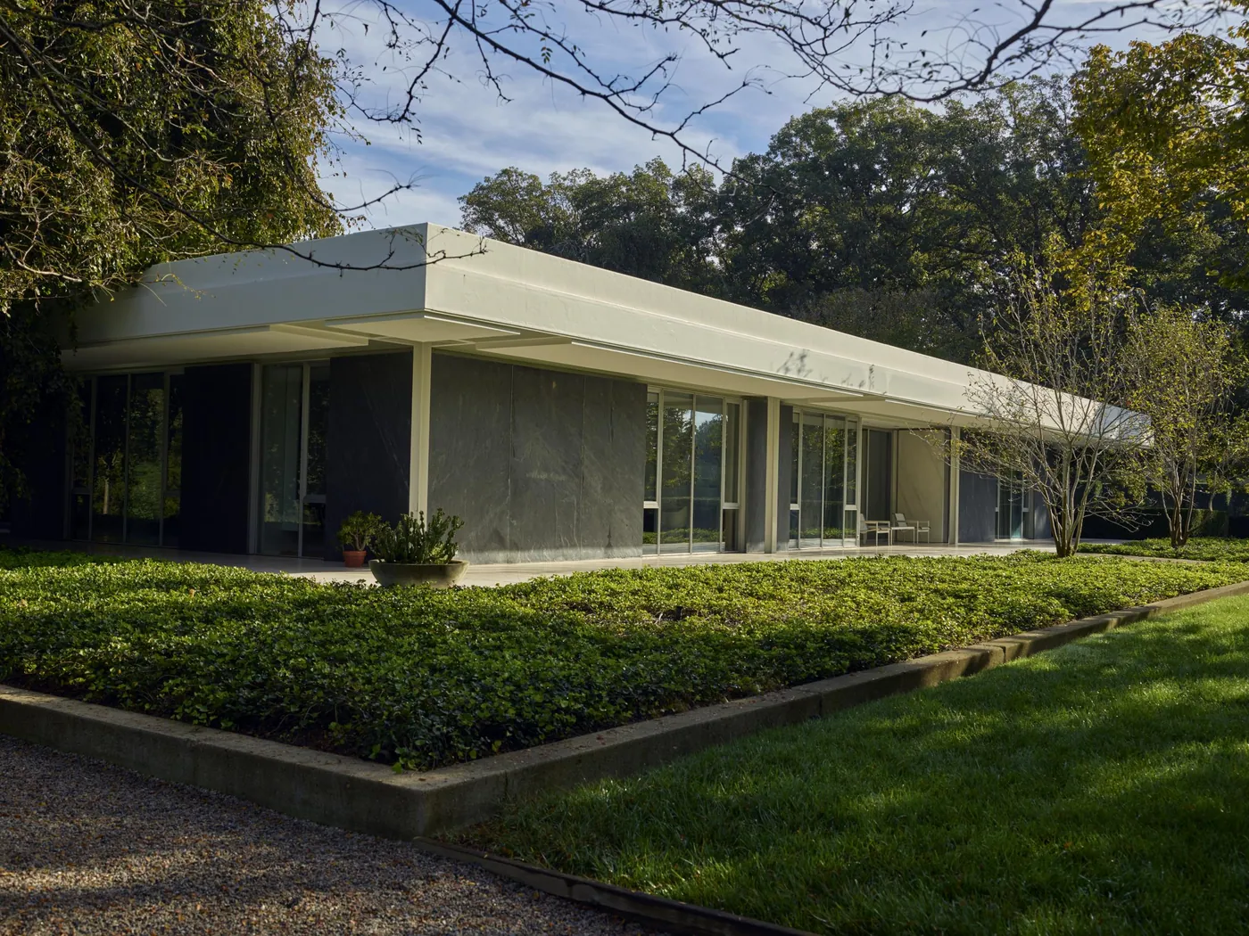A long, low 1957 modernist house with a flat white roof, glass walls and stone panels, set behind a clipped bed of ground cover and lawn