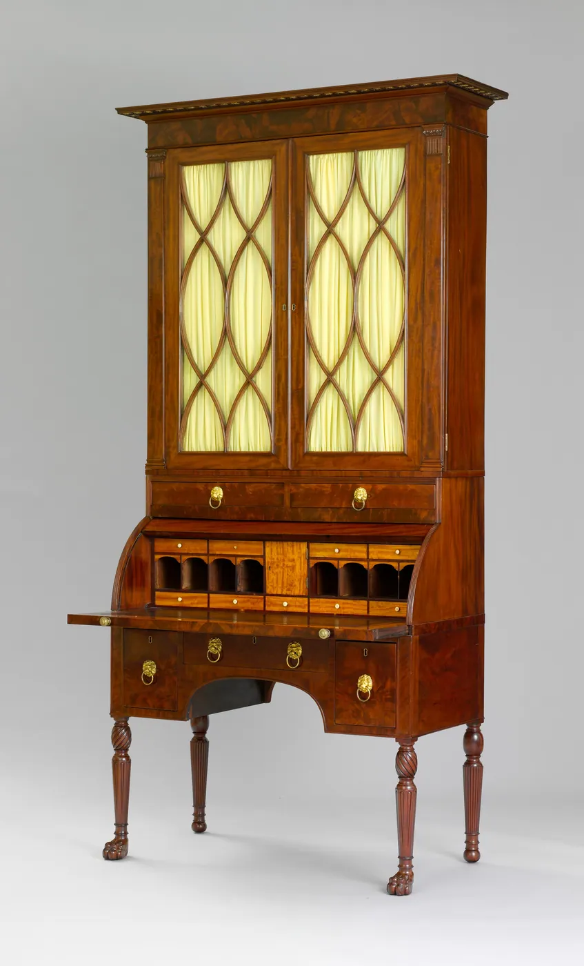 Mahogany cylinder desk with its writing surface pulled out, pigeonholes and small drawers, beneath a glazed bookcase with arched tracery doors